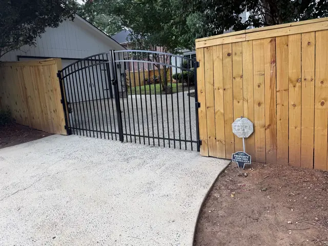 Black arched double drive gate between wood privacy fence sections at a concrete driveway.