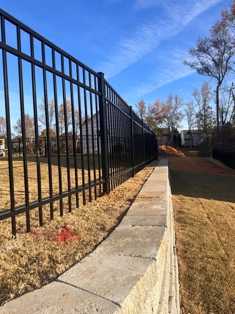 Black aluminum fence with three rails set behind a stone retaining wall.