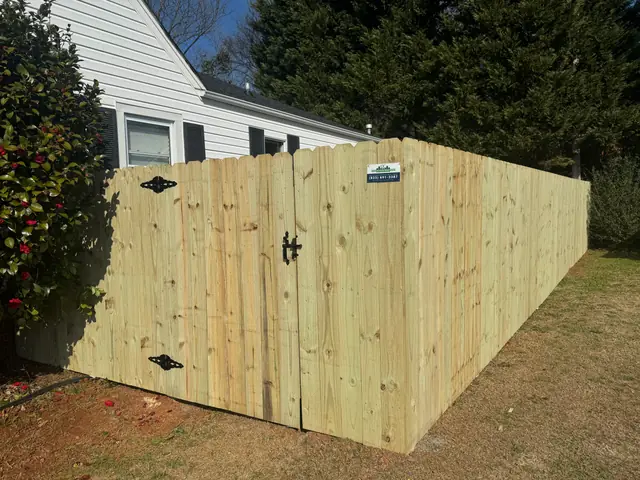 Dog-ear wood privacy fence with a gate and black hardware beside a white house.