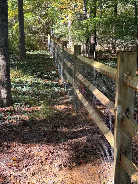 Long run of three-rail split rail fence with welded wire through a wooded backyard.