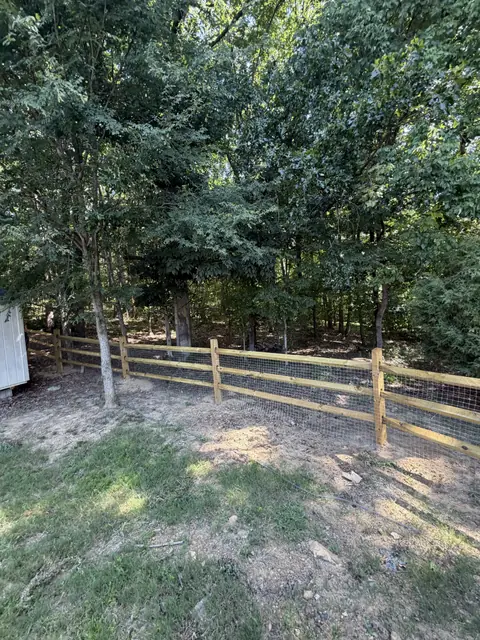 Three-rail split rail fence with welded wire at the edge of a wooded yard.